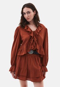 Blusa ruggine con balze e maniche lunghe, dotata di chiusura a cravatta sul collo. Abbinata a una gonna a strati coordinata e una cintura nera.