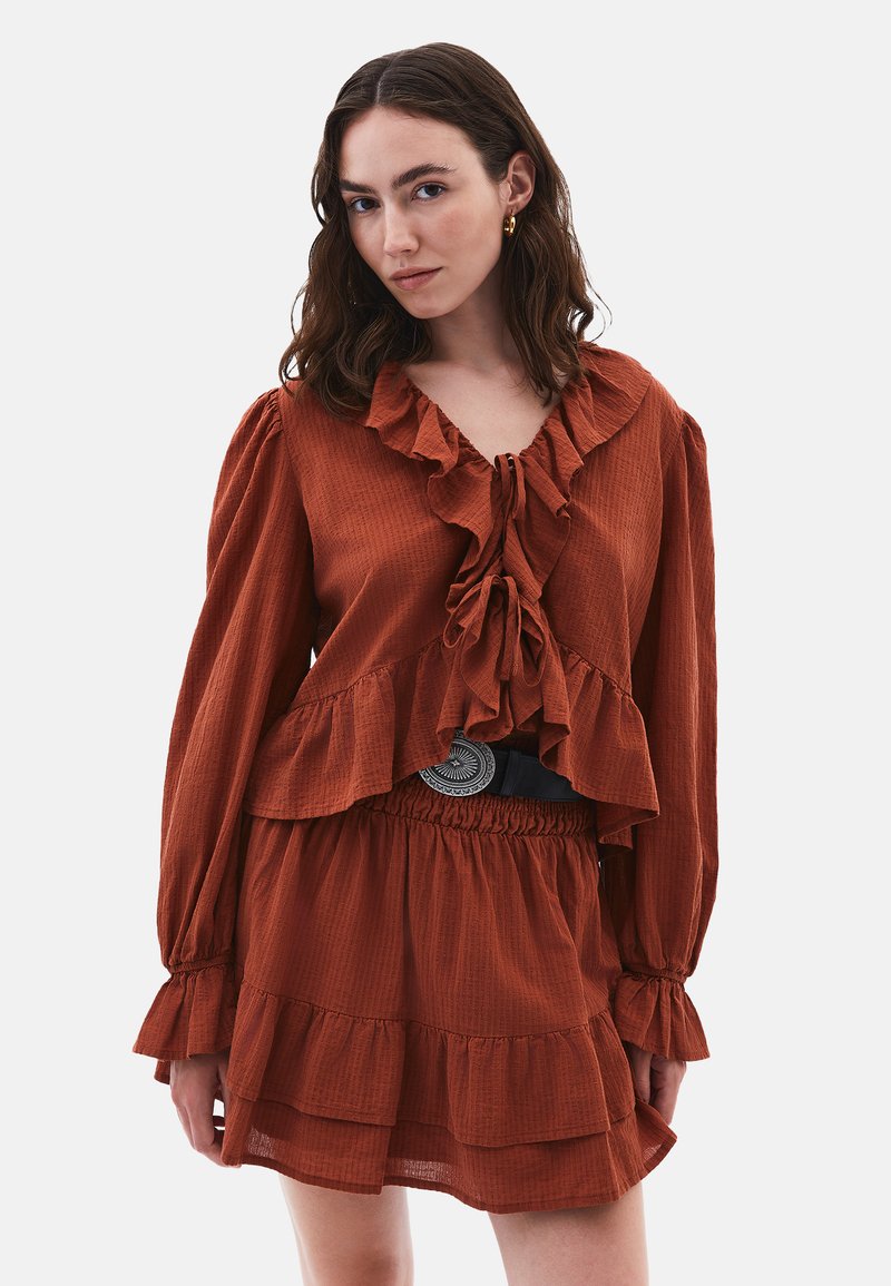 Blusa ruggine con balze e maniche lunghe, dotata di chiusura a cravatta sul collo. Abbinata a una gonna a strati coordinata e una cintura nera.