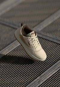 Lys beige sneakers med en teksturert overflate, mørkebrune detaljer, snøring og gummisåle, med en logo på tungen.