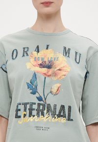 Lysegrønn bomulls T-skjorte med rund hals, med blomstertrykk i oransje og blått, og tekst i fet svart og gull.