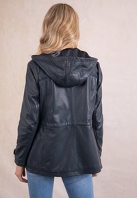 Blonde woman wearing a black leather jacket with hood, shown from the back, standing against a plain beige background.