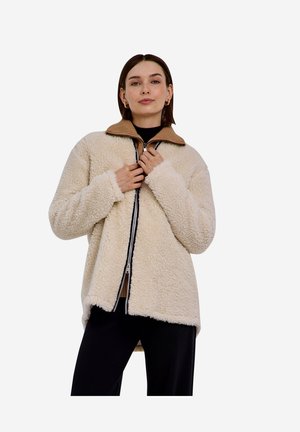 Junge Frau trägt eine beige Sherpa-Zip-up-Jacke über einem braunen Oberteil mit Kragen und schwarzen Hosen, steht vor einem einfachen weißen Hintergrund.