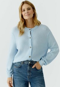 Frau mit hellblondem Haar trägt eine blassblaue, geknöpfte Strickjacke und hoch taillierte blaue Jeans, steht mit einer Hand in der Tasche.