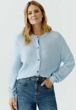 Woman with light blonde hair wears a pale blue buttoned cardigan and high-waisted blue jeans, standing with one hand in a pocket.