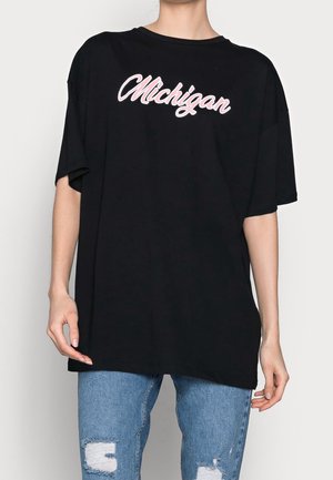 Personne portant un t-shirt noir ample avec "Michigan" en écriture blanche et rose, associé à un jean bleu clair déchiré.