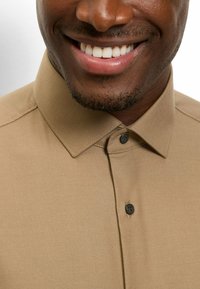 Hellbraunes Hemd mit Knöpfen und einem strukturierten Kragen, das eine einfarbige Textur und einen schwarzen Knopf aufweist. Nahaufnahme des Kragen- und Halsbereichs.