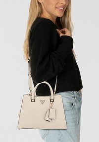 Donna in maglione nero e jeans, con una borsa Guess beige a manici superiori e tracolla sulla spalla.