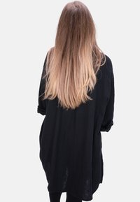 Person mit langen, glatten braunen und blonden Ombre-Haar, die ein lockeres schwarzes Langarmkleid trägt und mit dem Rücken zu einem weißen Hintergrund steht.