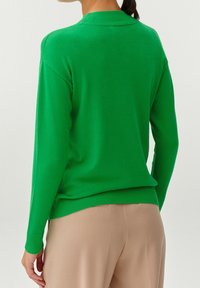Femme portant un pull vert clair à manches longues et un pantalon beige, vue de dos sur un fond uni.