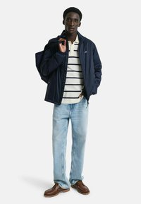 Marine lichtgewicht jack, gestreepte witte en marineblauwe polo, lichtblauwe jeans en bruine schoenen. Model poseert met een donkere schoudertas.