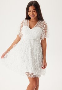 Kort vit klänning med en figurnära överdel och utsvängd kjol. Blommig broderi och genomskinliga, korta flygande ärmar förstärker designen.