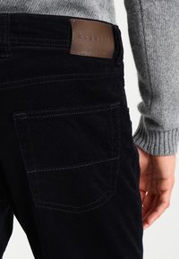 Tmavě modré manšestrové kalhoty s koženou nášivkou s nápisem "bugatti" v pase, s zadními kapsami a viditelnými detaily švů.