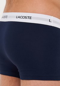 Tamno plave pamučne bokserice s bijelim elastičnim pojasom na kojem se nalazi logo "LACOSTE" i sivi pojas; glatka tekstura i prilagođen dizajn.