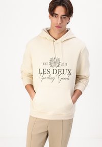 Cream-colored hooded sweatshirt with a front pocket and gray printed logo. Made of soft fabric, featuring ribbed cuffs and hem.