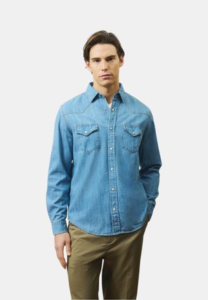 Giovane uomo che indossa una camicia di jeans azzurro chiaro con tasche sul petto e pantaloni kaki, in piedi davanti a uno sfondo neutro.