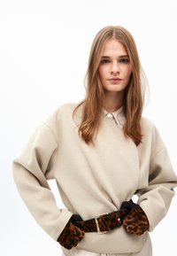 Beige fleece neule, jossa on pyöristetyt olkapäät, vyötärölle kiinnitetty mustalla vyöllä, jossa on kultainen solki, yhdistettynä ruskeisiin leopardikuvioisiin käsineisiin.
