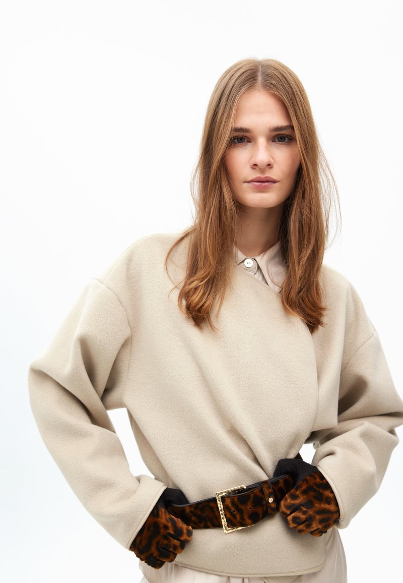 Beige fleece neule, jossa on pyöristetyt olkapäät, vyötärölle kiinnitetty mustalla vyöllä, jossa on kultainen solki, yhdistettynä ruskeisiin leopardikuvioisiin käsineisiin.
