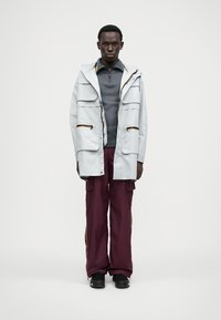 Veste imperméable gris clair avec plusieurs poches, associée à un haut gris foncé et un pantalon cargo bordeaux avec des bandes latérales, ainsi que des chaussures noires.