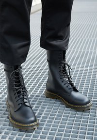 Botas de cuero negro con cordones y caña alta, costuras amarillas y suelas de goma gruesas. Textura suave y punta redondeada.