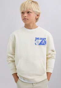 Sudadera de color crema con un estampado gráfico azul que presenta un baloncesto y una calavera. Tiene un cuello redondo y un bolsillo delantero.