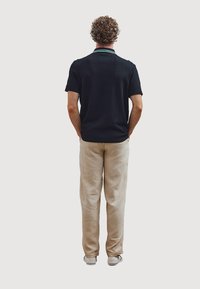 Zwarte polo met korte mouwen en een gestructureerde afwerking, voorzien van een groen-gestreepte kraag, gecombineerd met lichtbeige broeken en witte sneakers.