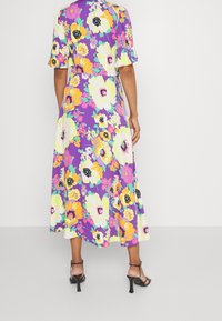 Donna che indossa un vestito midi viola con grandi fantasie floreali gialle, bianche e rosa, abbinato a sandali neri con tacchi a spillo e cinturini.