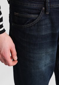 Jeans de mezclilla azul oscuro con un acabado ligeramente deslucido, que presentan un diseño clásico de cinco bolsillos y costuras amarillas en las costuras.