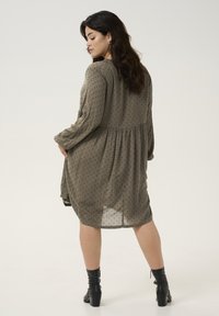 Robe vert olive, jusqu'aux genoux, à manches longues, avec un motif à pois, taille froncée et tissu léger et semi-transparent. Portée avec des bottines noires.