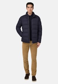 Tumes puffer-jakk ülikõrge kaelusega, horisontaalsete tikkimismustritega ja esi-zippiga. Paaritatud tan värvi korteriga pükstega ja tumedate jalanõudega.