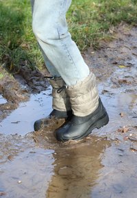 Schwarze wasserdichte Stiefel mit strukturierten Gummisohlen, kombiniert mit grauen isolierten Manschetten, stehend in einer schlammigen Pfütze.