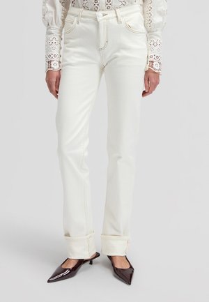 Donna che indossa jeans bianchi a gamba dritta con risvolti, una camicetta bianca in pizzo a maniche lunghe e décolleté nere con punta appuntita e tacco basso.