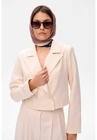 Lichtbeige dubbelzijdige cropped blazer met twee knopen, gecombineerd met een geplooide rok. Het model draagt zwarte zonnebrillen en een gestreepte sjaal.