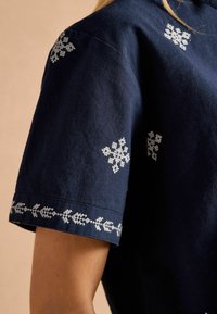 Cămașă albastră închis cu broderie albă având modele florale și geometrice, mâneci scurte și o croială lejeră. Detaliu de țesătură vizibil.