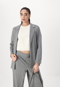 Gray checked blazer with a relaxed fit and notched lapels, worn over a textured cream top, paired with tailored gray pants.