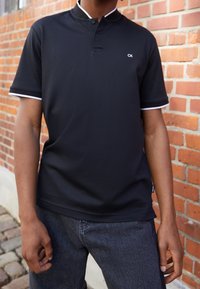 Polo noir avec col à boutons et bordure blanche contrastante aux manches. Confectionné en tissu lisse et respirant. Petit logo "CK" sur la poitrine.