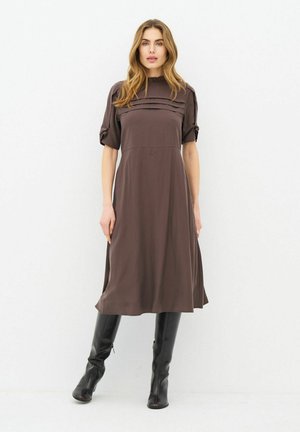 Femme aux longs cheveux châtain clair portant une robe midi marron avec un détail plissé et des bottes noires montantes, debout devant un fond blanc.