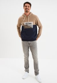 Felpa con cappuccio beige e blu navy, con tasca frontale e design grafico. Abbinata a jeans grigi e sneakers bianche.