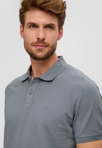 Polo gris avec un col classique, des manches courtes et un motif texturé subtil, arborant un logo sur le côté gauche.