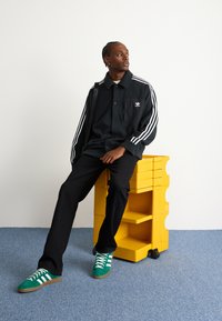 Černá košile na knoflíky s bílými pruhy, černé kalhoty a zelené tenisky s bílými akcenty, sedící na žluté pojízdné úložné jednotce.