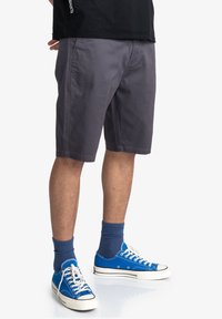 Pantaloni corti grigi in cotone con tasche laterali, abbinati a sneaker alte blu e calze navy. Design semplice, vestibilità fino al ginocchio.