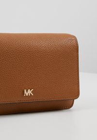 Portefeuille en cuir brun texturé avec un logo MK doré sur le rabat avant, sur un fond gris clair uni.