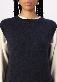 Colete de malha azul marinho com um acabamento texturizado, gola redonda, e sobreposto a uma blusa de manga longa creme.