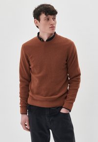 Suéter marrón texturizado con un cuello redondo, usado sobre una camisa de cuello negro, combinado con jeans oscuros. Diseño simple, con puños y dobladillo acanalados.