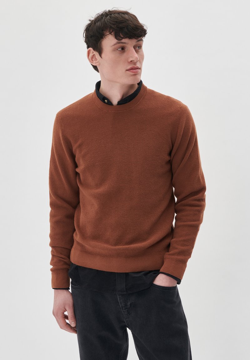 Suéter marrón texturizado con un cuello redondo, usado sobre una camisa de cuello negro, combinado con jeans oscuros. Diseño simple, con puños y dobladillo acanalados.