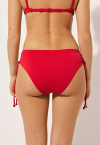 Röda bikiniunderdelar med justerbara sidoremmar. Slät yta, medelhögt skydd och en klassisk design. Bakifrån som visar passform och stil.