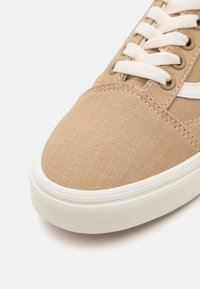 Beige kanvas sneakers med hvite snørebånd, en hvit side stripe og en teksturert gummisåle. Nærbilde av tåområdet og det øvre materialet.