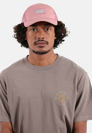 Homme aux cheveux bouclés portant une casquette rose Oxbow et un t-shirt taupe avec un logo soleil jaune et le texte Oxbow sur la poitrine.