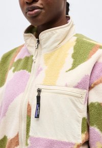 Κοντινό πλάνο ενός ατόμου που φοράει ζακέτα από απαλό υλικό με παστέλ καμουφλάζ, μπεζ γιακά και τσέπη στο στήθος με φερμουάρ που φέρει το λογότυπο της Patagonia.