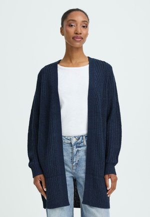 Mörkblå stickad cardigan med öppen front, strukturerad mönster, långa ärmar och ribbade muddar, bärs över en vit skjorta och denimjeans.