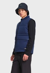 Homme portant une veste matelassée bleu marine par-dessus une chemise noire à manches longues, un pantalon noir et un bob noir avec le logo "AIGLE".